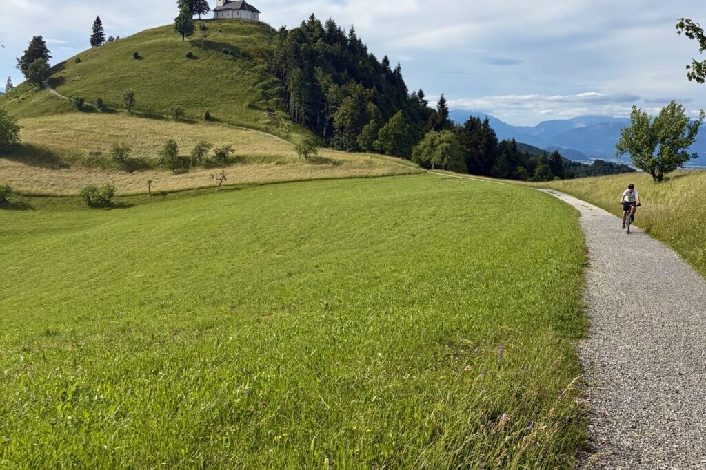 Gravel cyclist riding a gravel path through green meadows below a hilltop church, with the Alps on the horizon