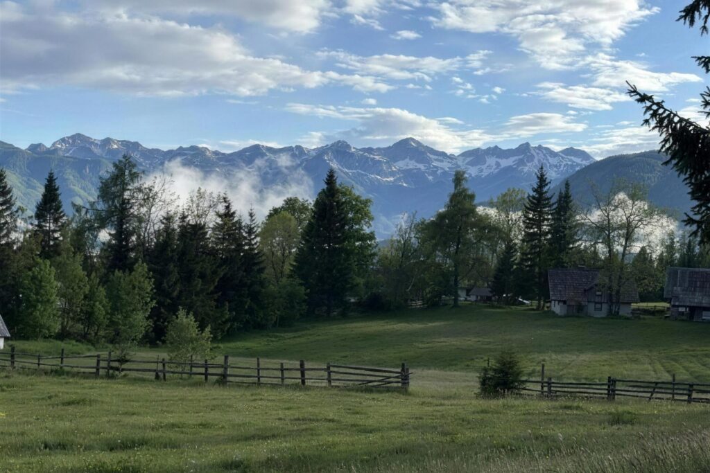 A fenced meadow and traditional wooden farm buildings below the snow-dusted Julian Alps